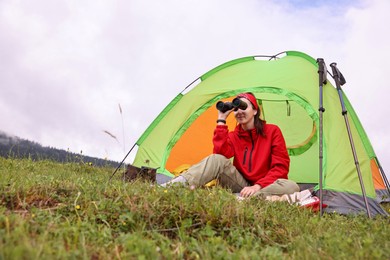 Photo of Young woman sitting in tent and looking through binoculars on green grass outdoors, low angle view