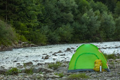 Camping tent, backpack and thermo bottle on stones near river in mountains