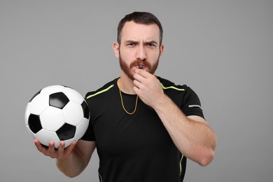 Photo of Young man with soccer ball blowing whistle on grey background