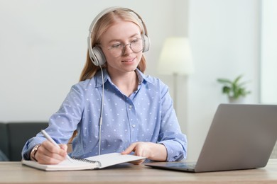 Photo of Interpreter in headphones taking notes while working with laptop at table indoors