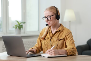 Interpreter in headset taking notes while working with laptop at table indoors