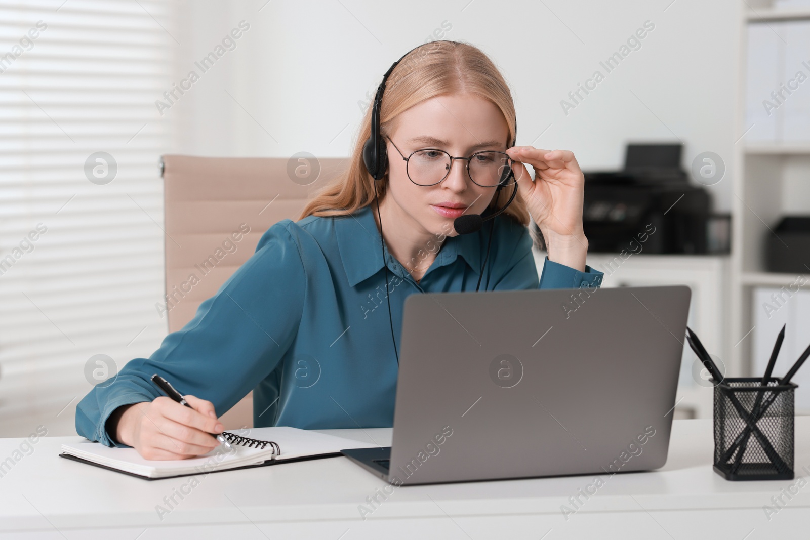 Photo of Interpreter in headset taking notes while working with laptop at table indoors