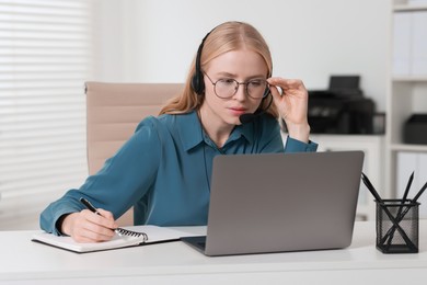 Interpreter in headset taking notes while working with laptop at table indoors