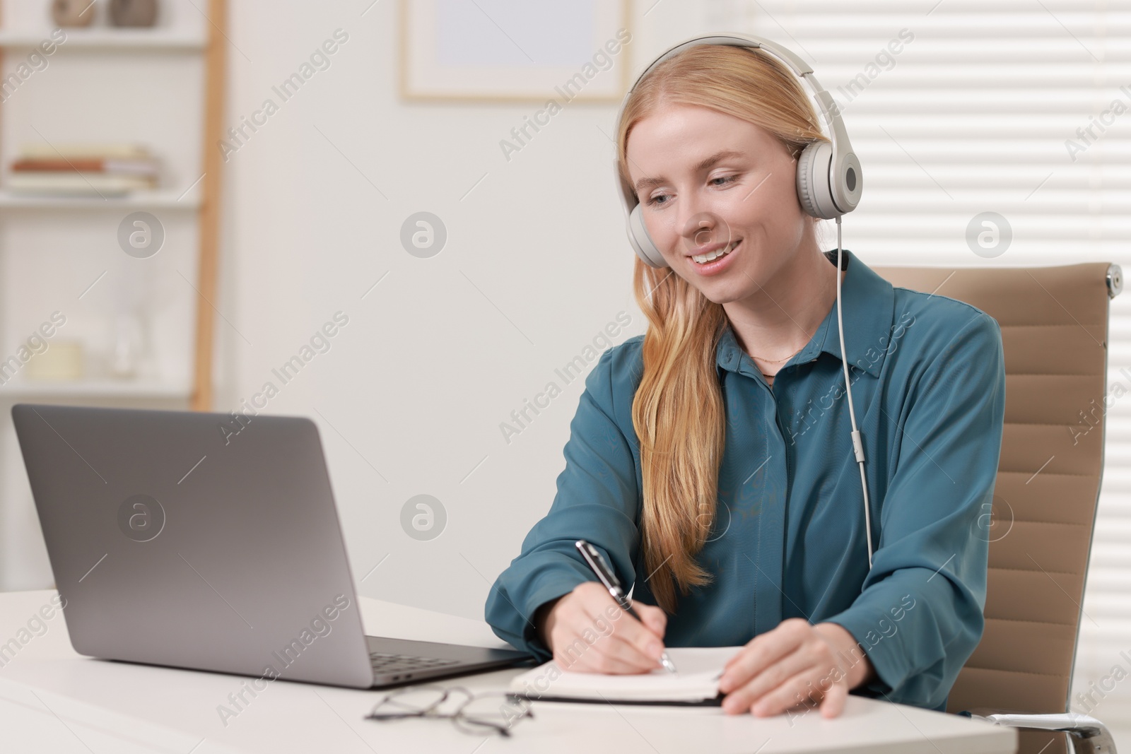 Photo of Interpreter in headphones taking notes while working with laptop at table indoors