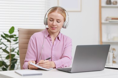 Interpreter in headphones taking notes while having video chat via laptop at table indoors