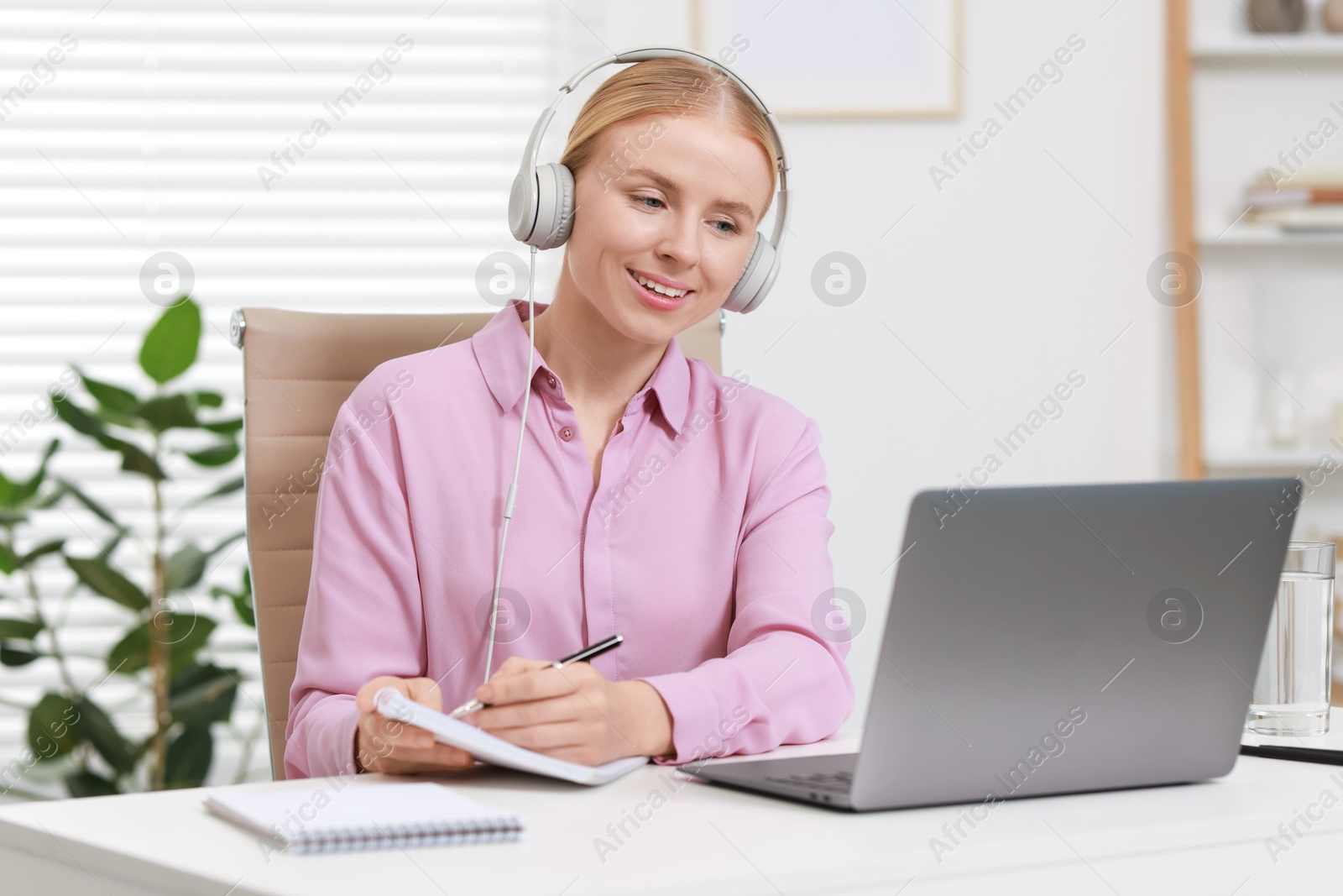 Photo of Interpreter in headphones taking notes while having video chat via laptop at table indoors