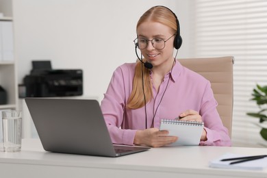 Interpreter in headset taking notes while working with laptop at table indoors