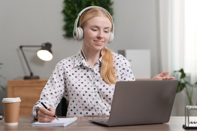 Interpreter in headphones taking notes while having video chat via laptop at table indoors