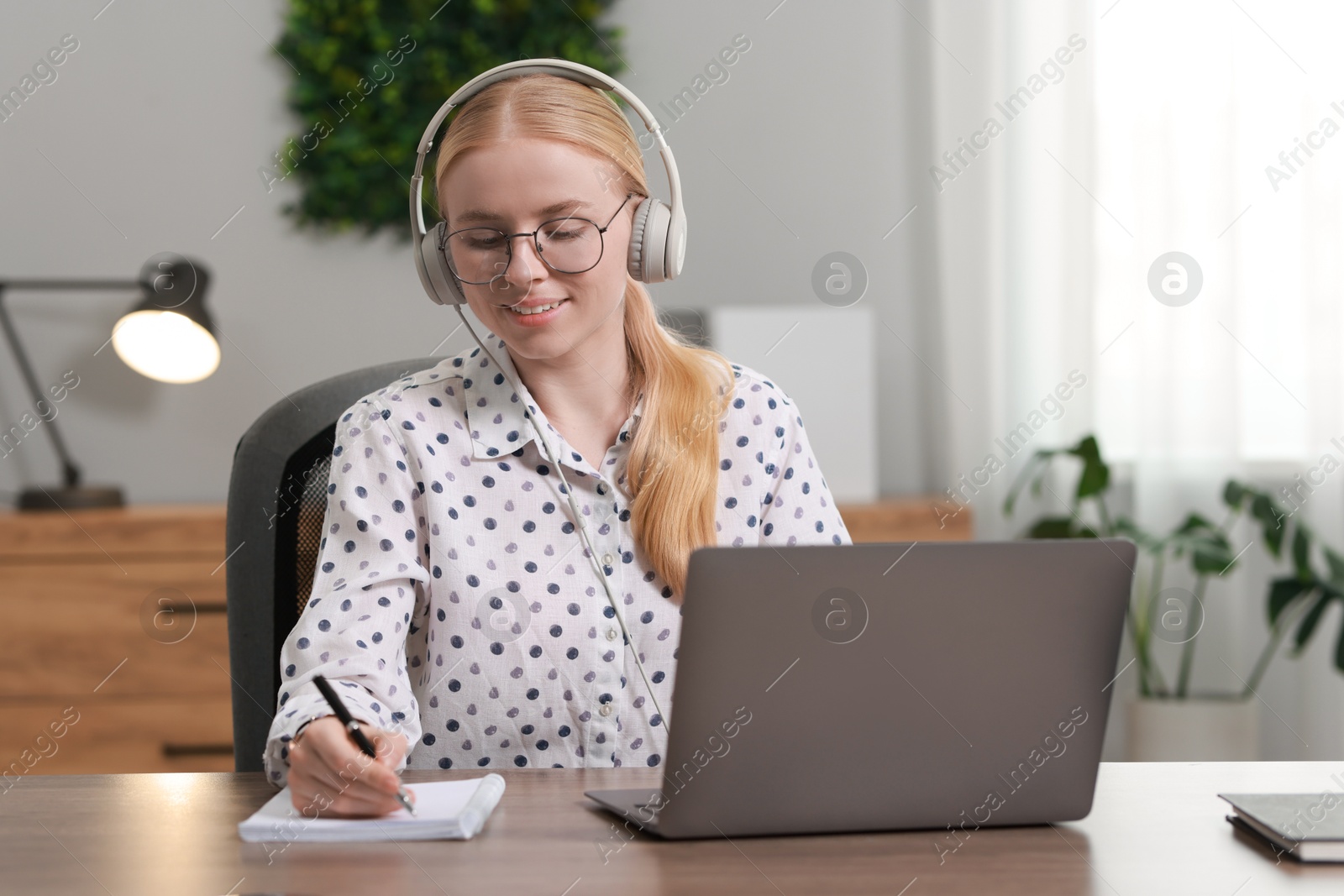 Photo of Interpreter in headphones taking notes while working with laptop at table indoors