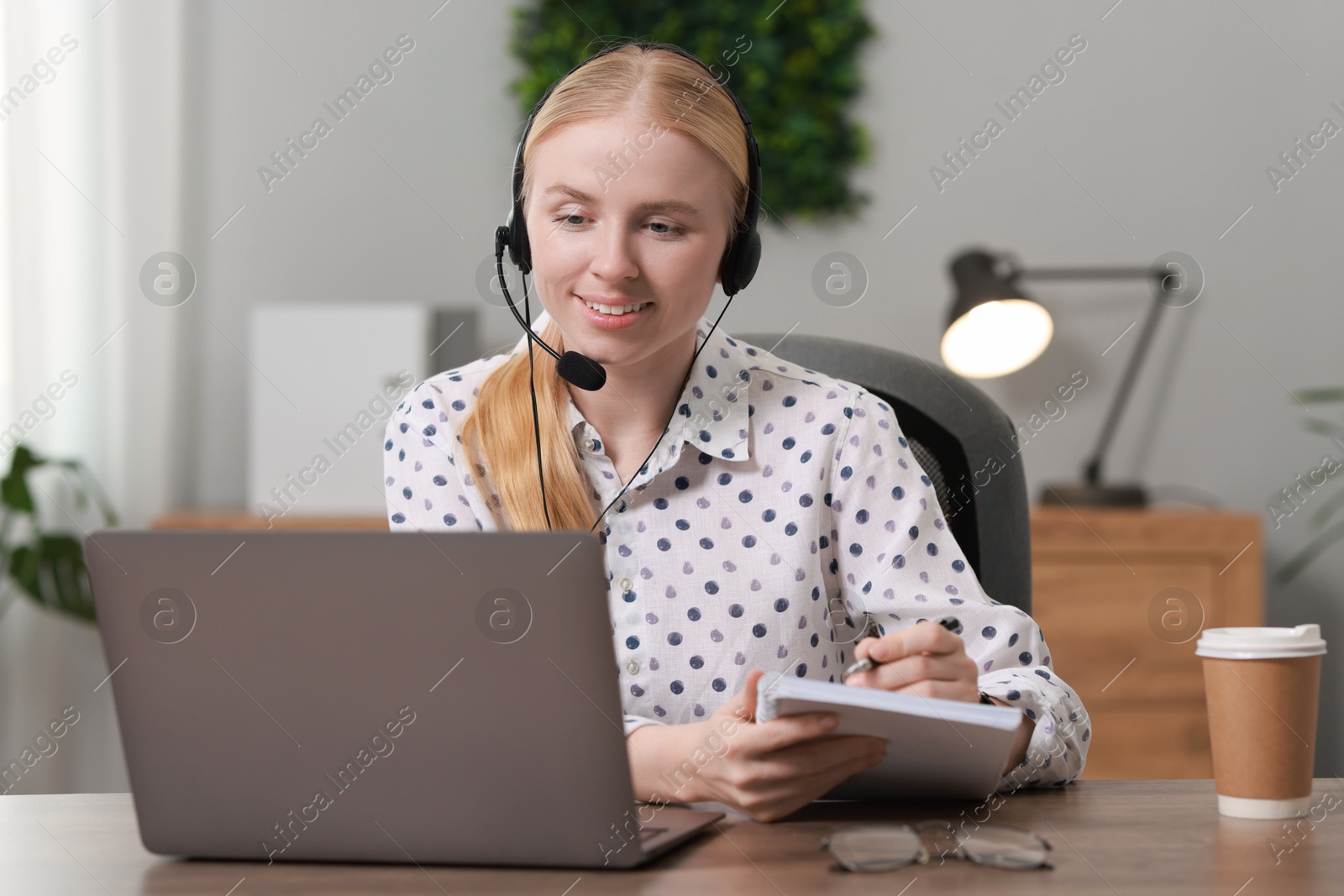 Photo of Interpreter in headset taking notes while working with laptop at table indoors