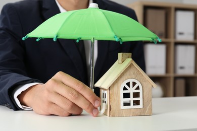 Photo of Real estate insurance. Man holding small umbrella above wooden house figure at light table, closeup