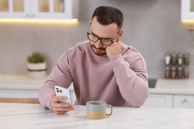 Handsome man using smartphone at white table in kitchen