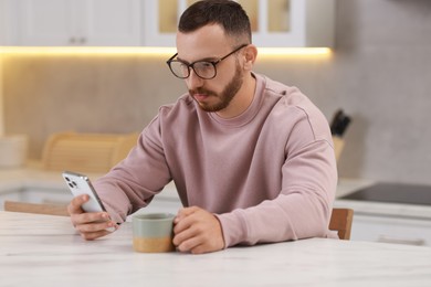 Handsome man using smartphone at white table in kitchen