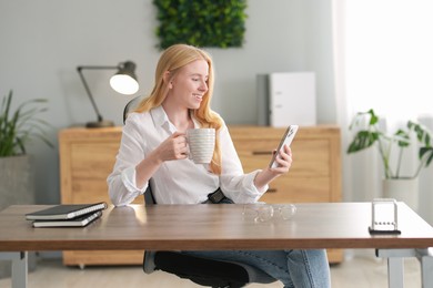 Photo of Smiling woman using smartphone and drinking coffee at table in office