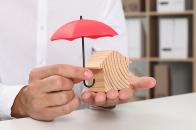 Photo of Real estate insurance. Man holding small umbrella above wooden house figure at light table, closeup