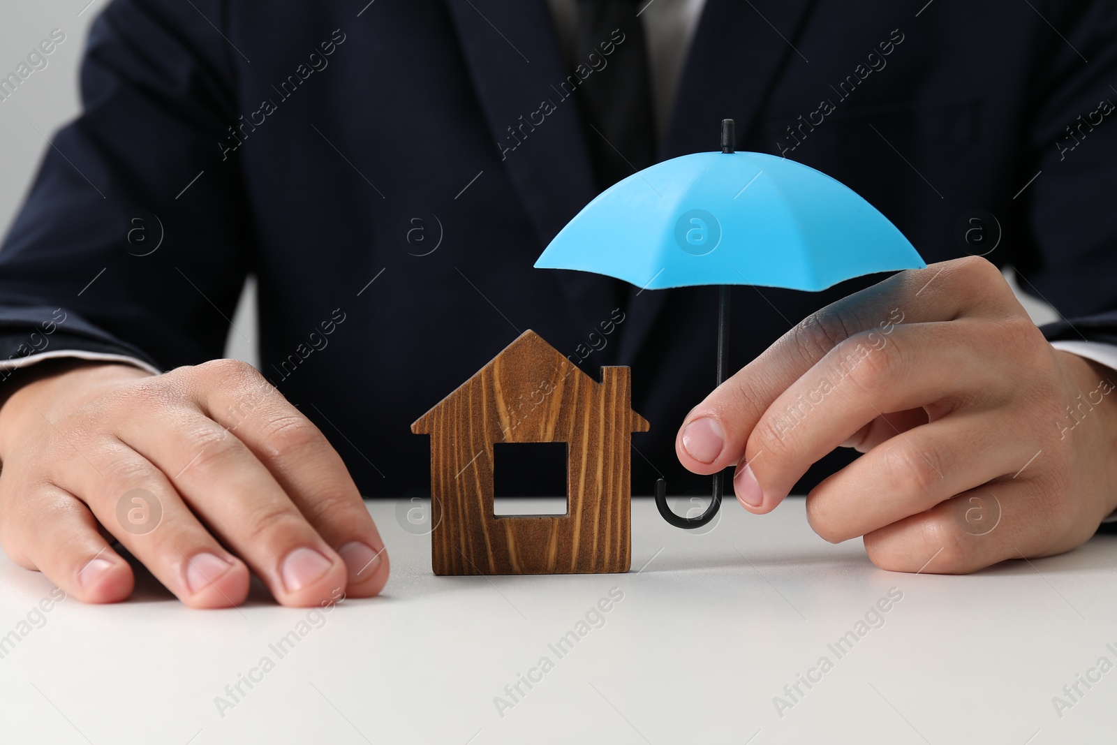 Photo of Real estate insurance. Man holding small umbrella above wooden house figure at light table, closeup