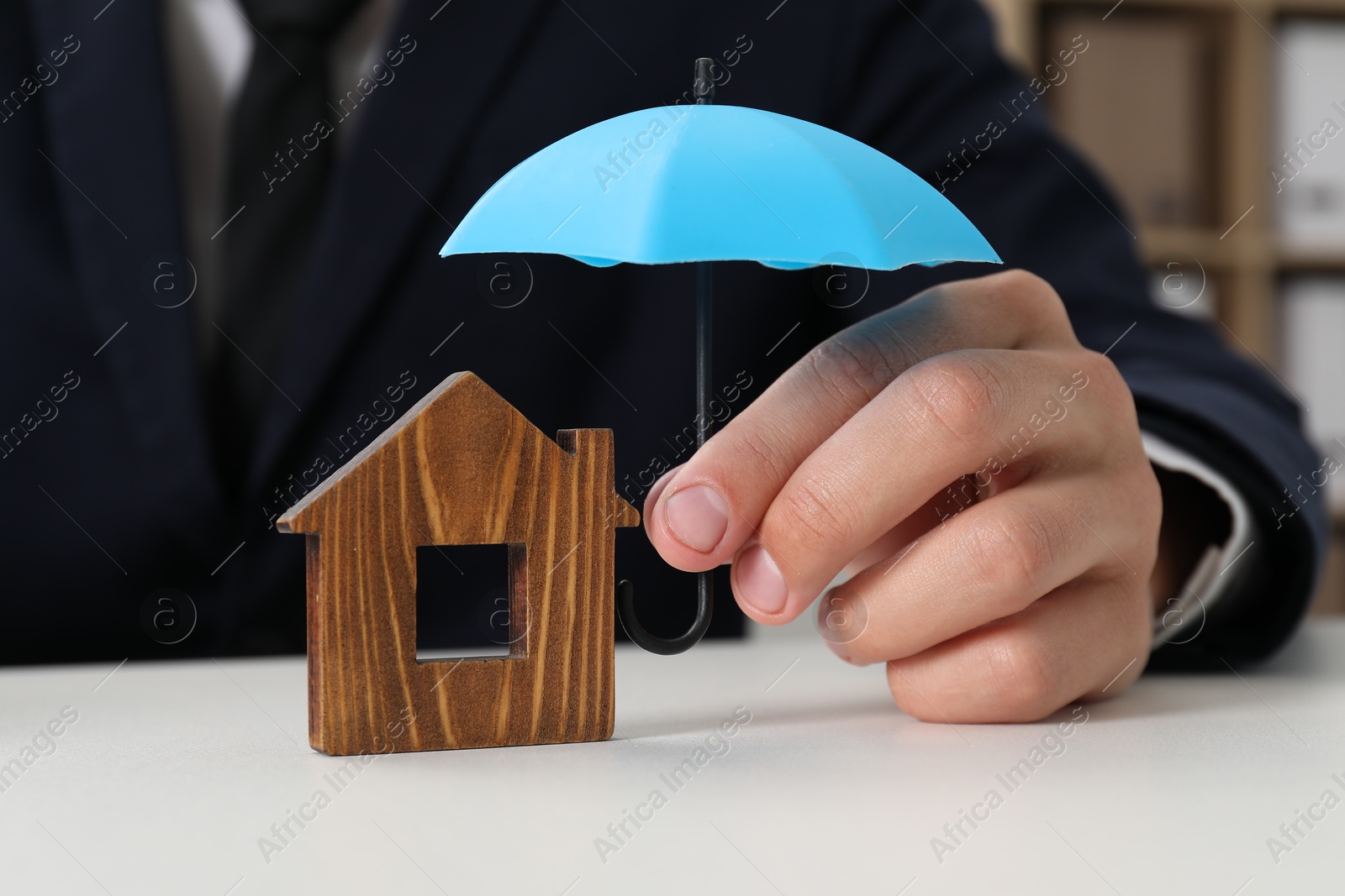 Photo of Real estate insurance. Man holding small umbrella above wooden house figure at light table, closeup