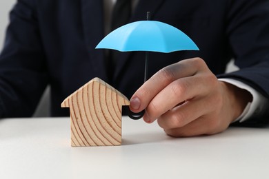 Photo of Real estate insurance. Man holding small umbrella above wooden house figure at light table, closeup