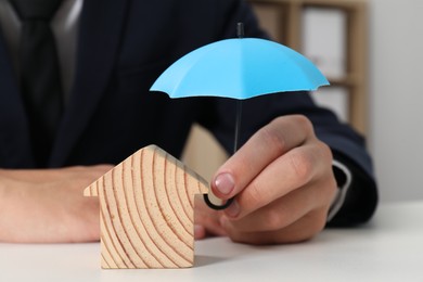 Photo of Real estate insurance. Man holding small umbrella above wooden house figure at light table, closeup