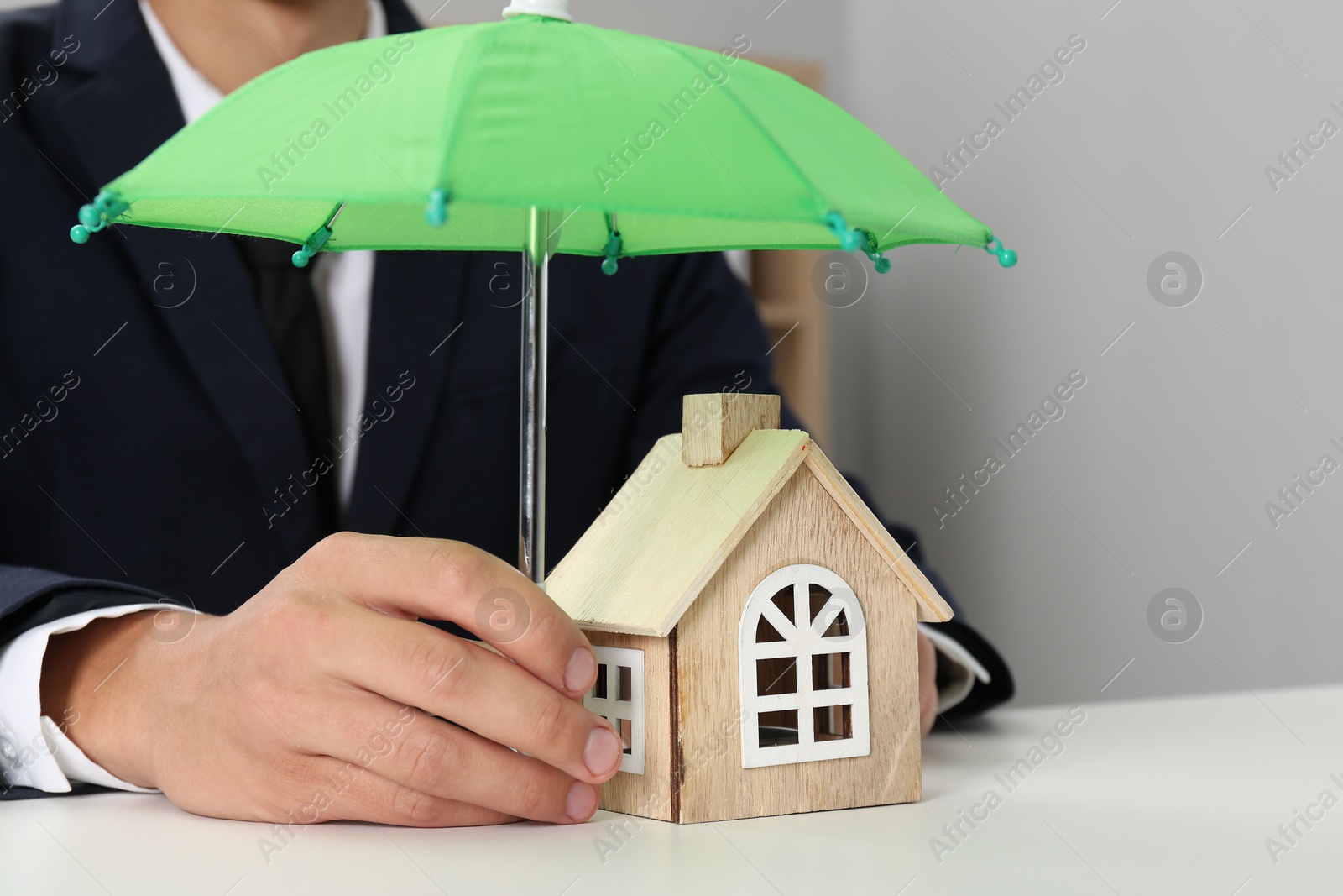 Photo of Real estate insurance. Man holding small umbrella above wooden house figure at light table, closeup