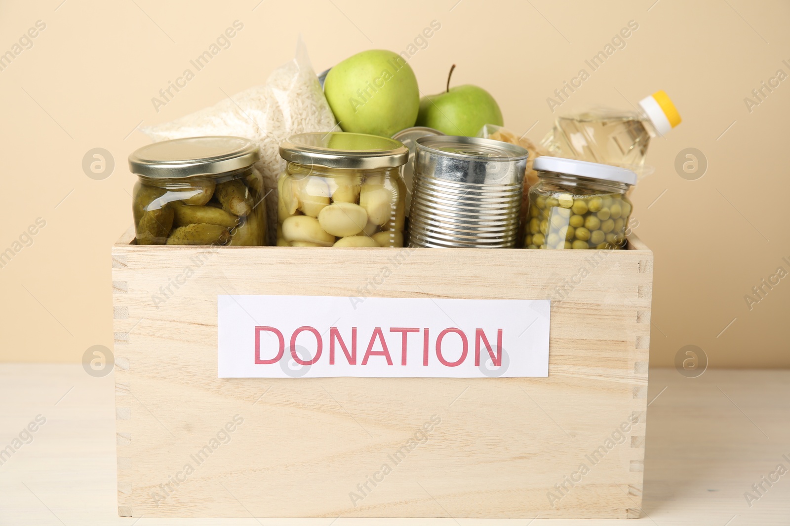 Photo of Different food products for donation in wooden crate on table against beige background