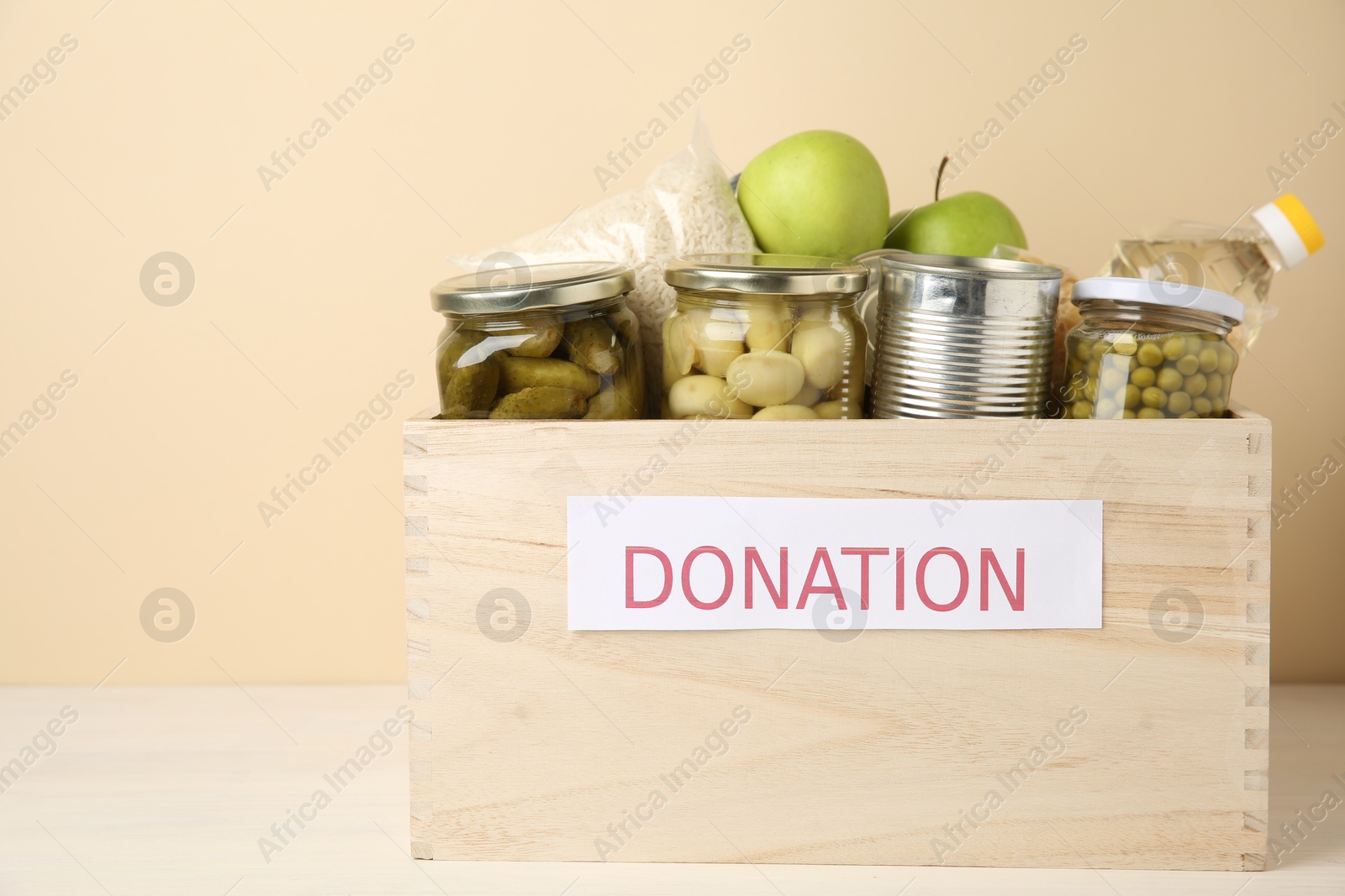 Photo of Different food products for donation in wooden crate on table against beige background