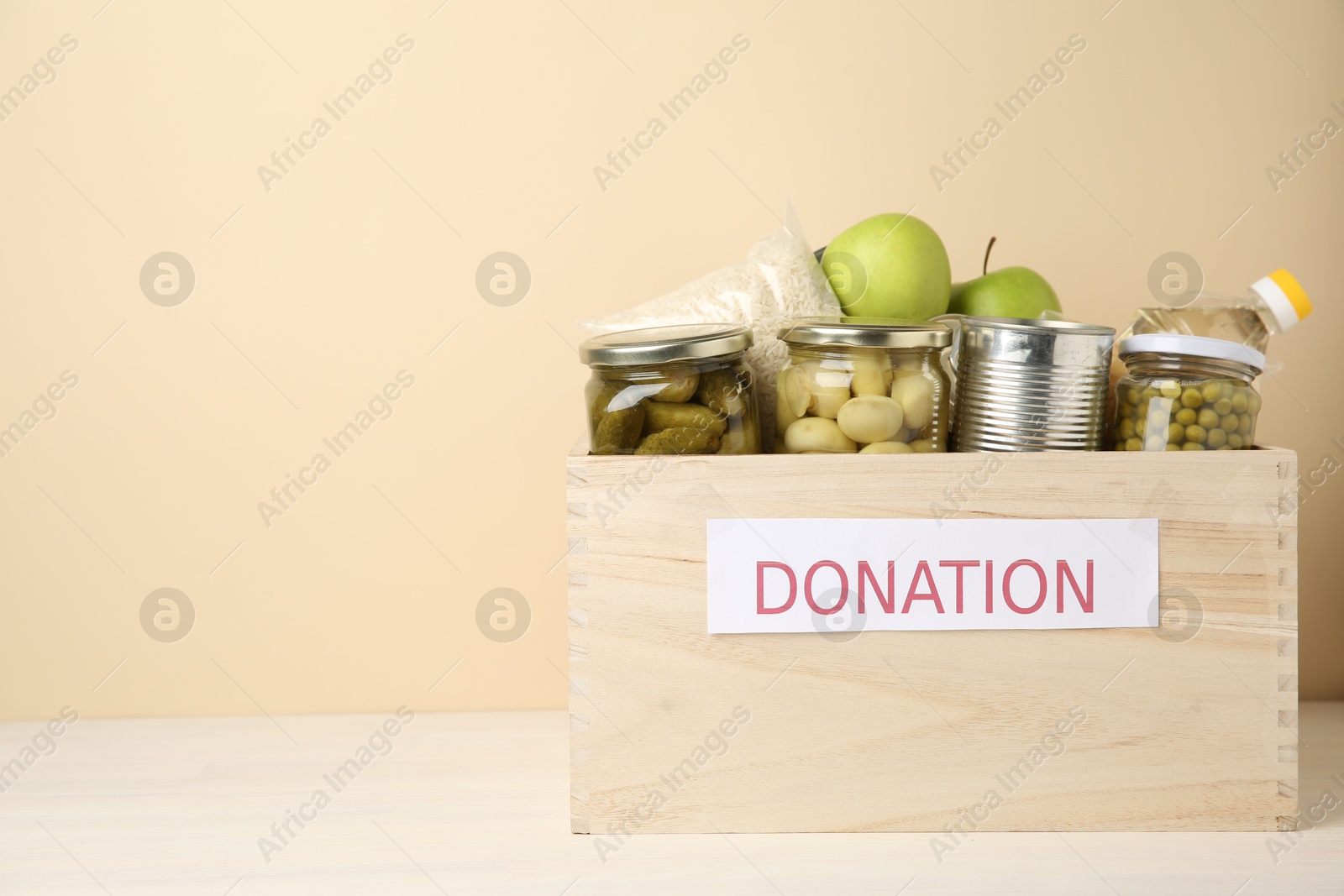 Photo of Different food products for donation in wooden crate on table against beige background, space for text