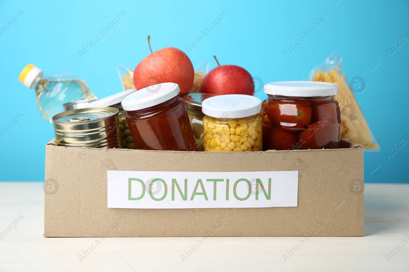 Photo of Different food products for donation in box on table against light blue background