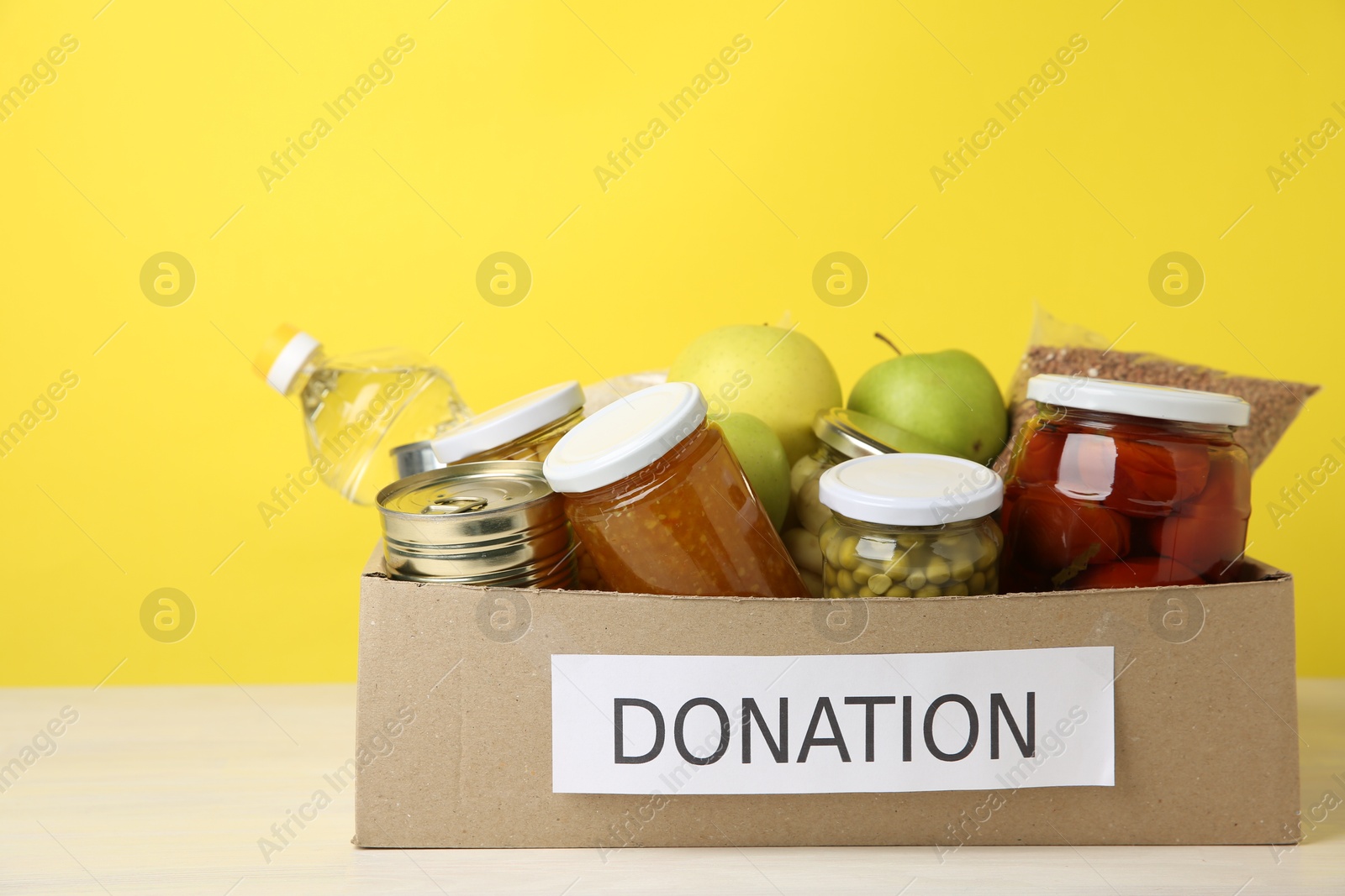 Photo of Different food products for donation in box on table against yellow background