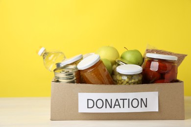 Photo of Different food products for donation in box on table against yellow background