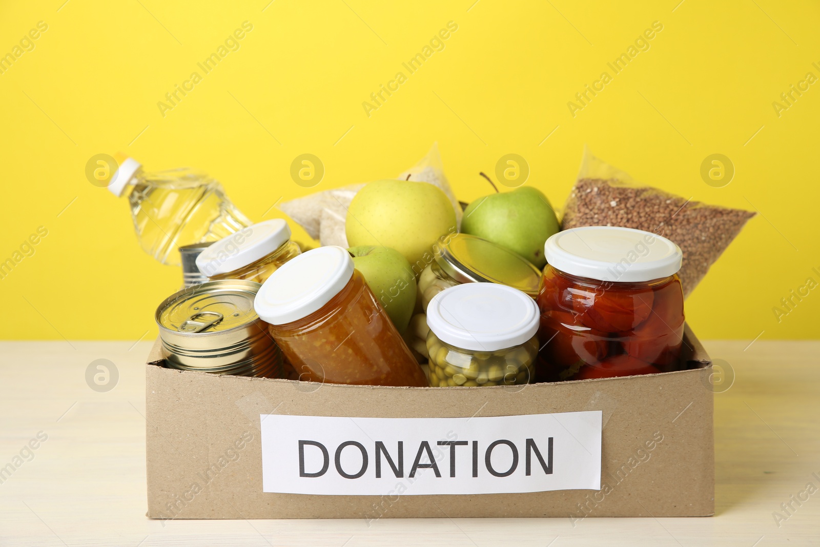 Photo of Different food products for donation in box on table against yellow background