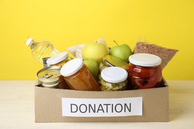 Photo of Different food products for donation in box on table against yellow background