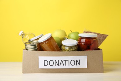 Photo of Different food products for donation in box on table against yellow background