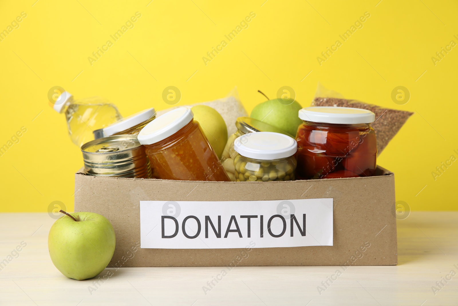 Photo of Different food products for donation in box on table against yellow background