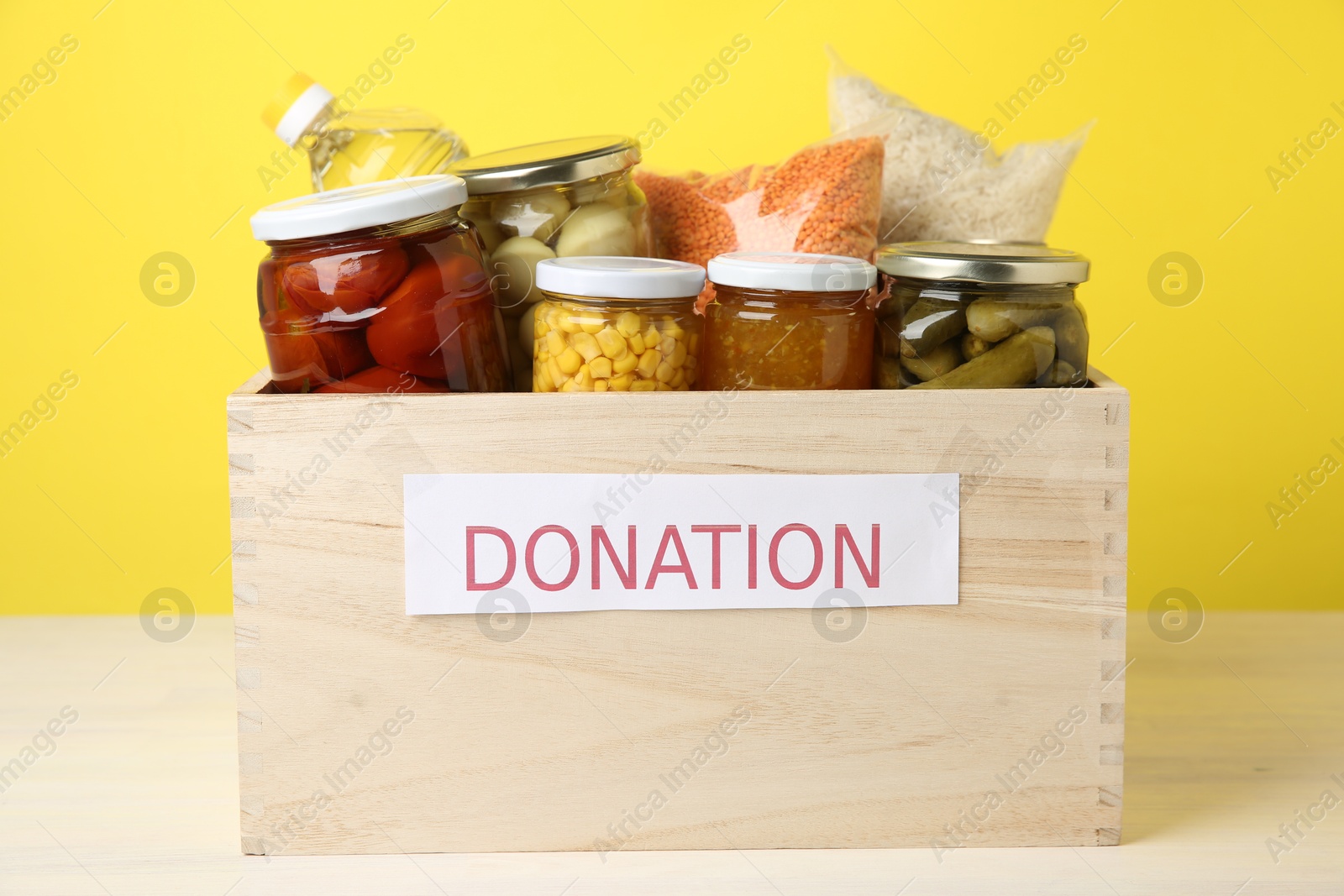 Photo of Different food products for donation in wooden crate on table against yellow background