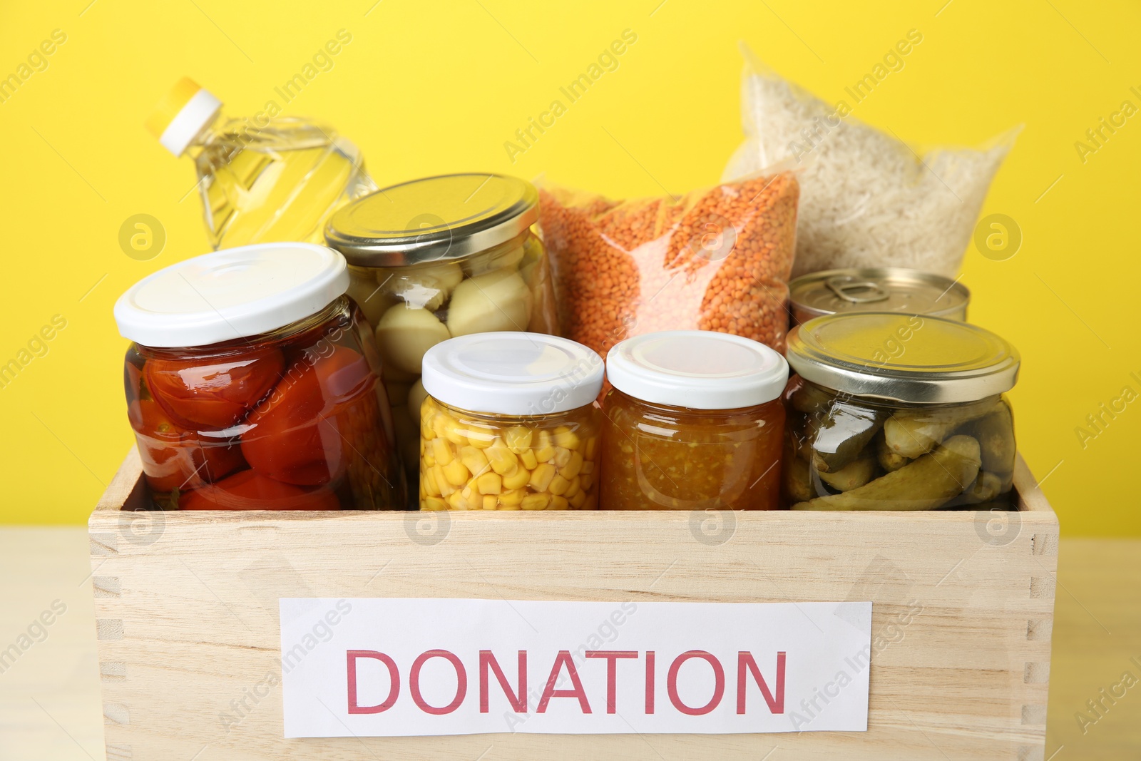 Photo of Different food products for donation in wooden crate on table against yellow background, closeup