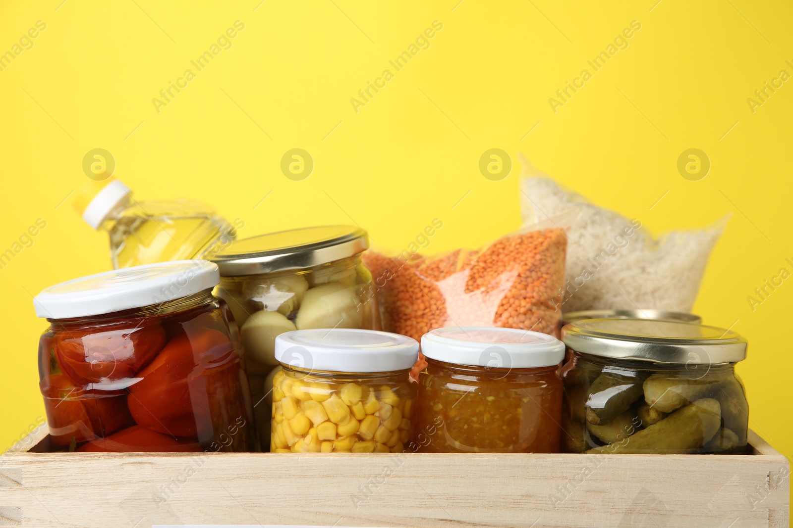 Photo of Different food products for donation in wooden crate on yellow background, closeup