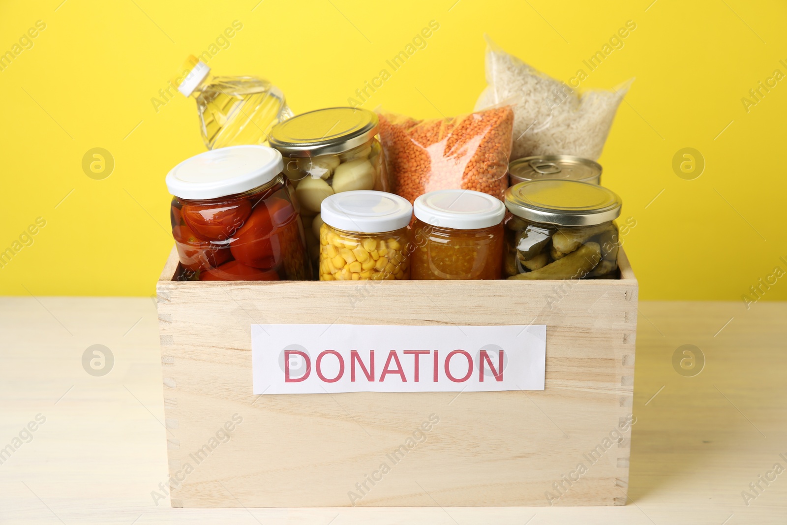 Photo of Different food products for donation in wooden crate on table against yellow background