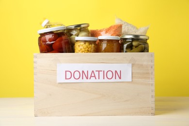 Photo of Different food products for donation in wooden crate on table against yellow background