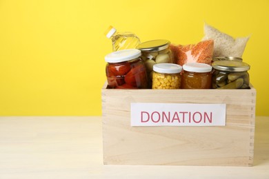 Photo of Different food products for donation in wooden crate on table against yellow background, space for text