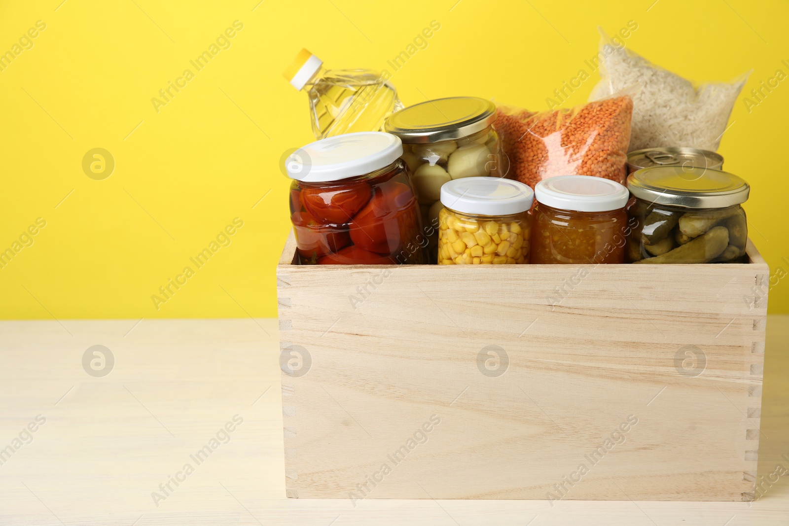 Photo of Different food products for donation in wooden crate on table against yellow background, space for text