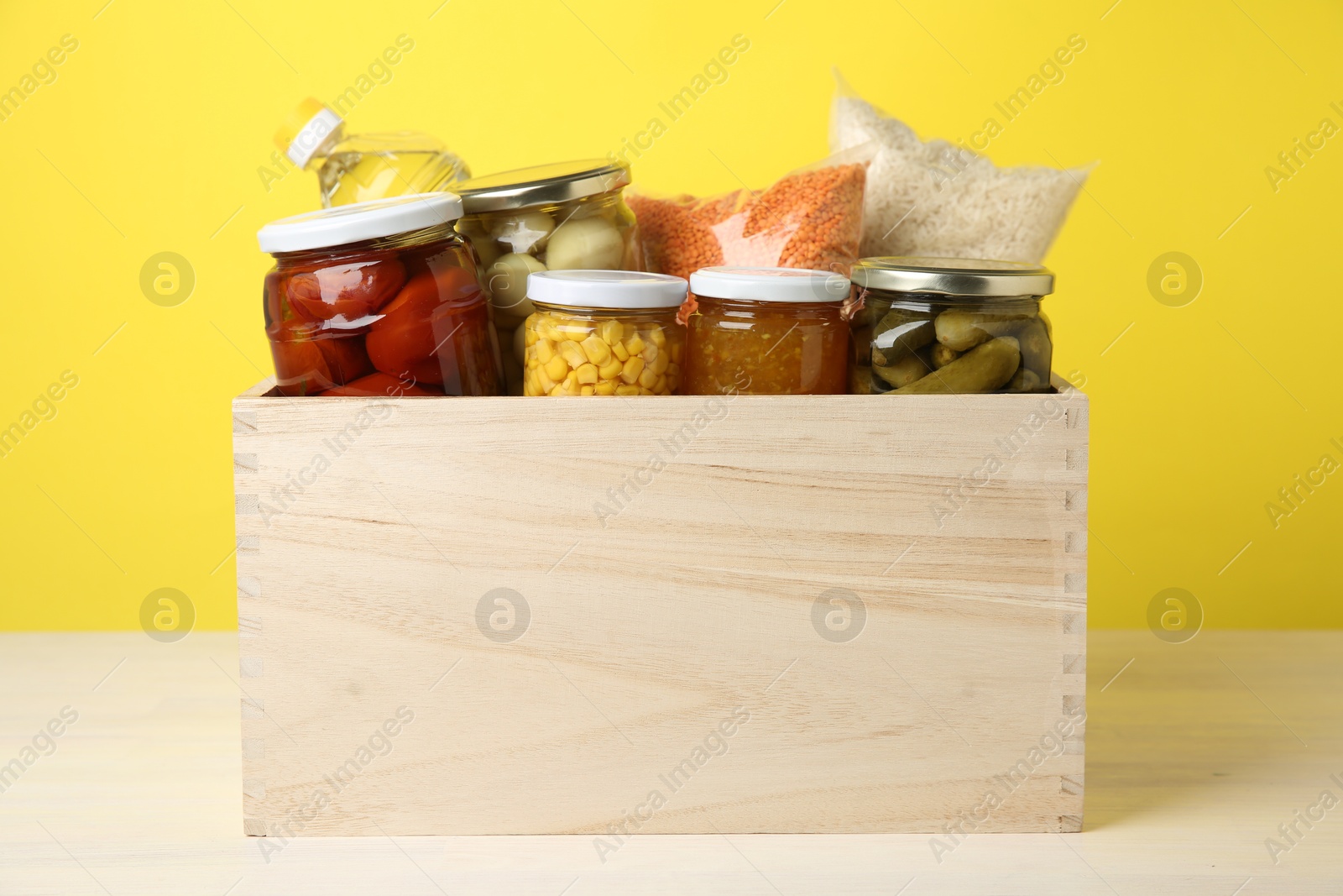 Photo of Different food products for donation in wooden crate on table against yellow background