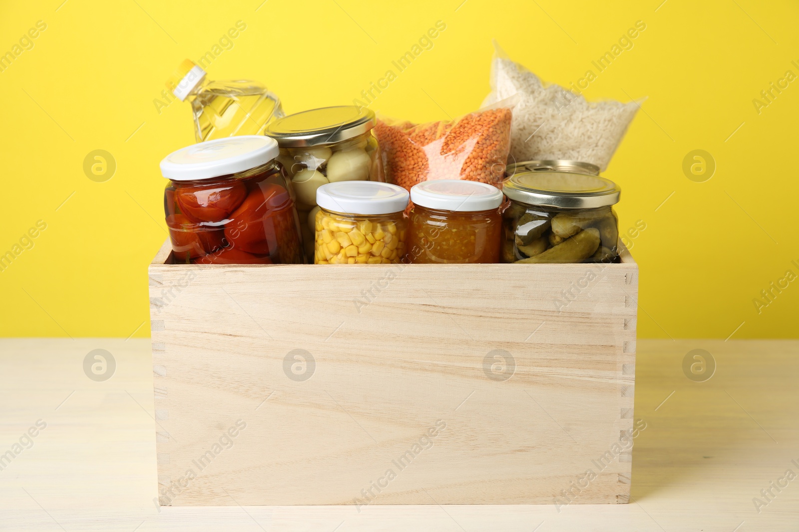 Photo of Different food products for donation in wooden crate on table against yellow background