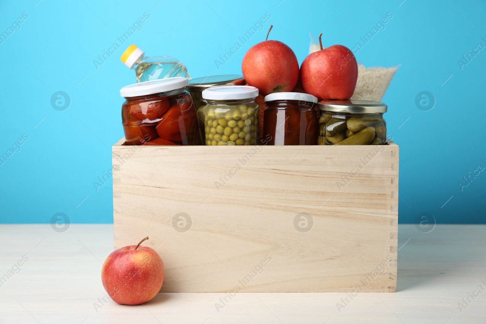 Photo of Different food products for donation in wooden crate on table against light blue background