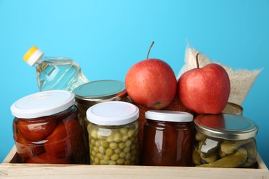 Photo of Different food products for donation in wooden crate on light blue background, closeup