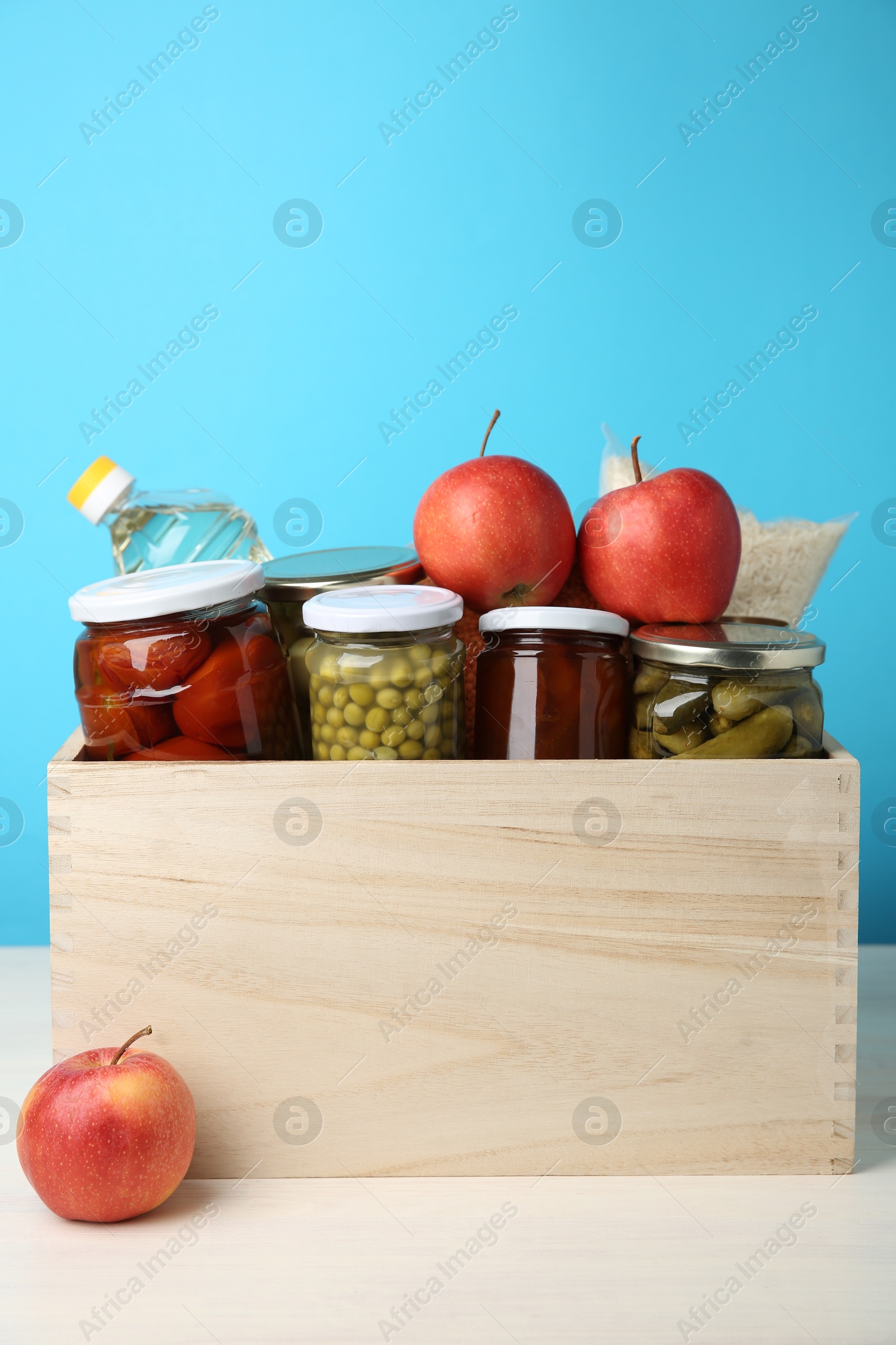 Photo of Different food products for donation in wooden crate on table against light blue background