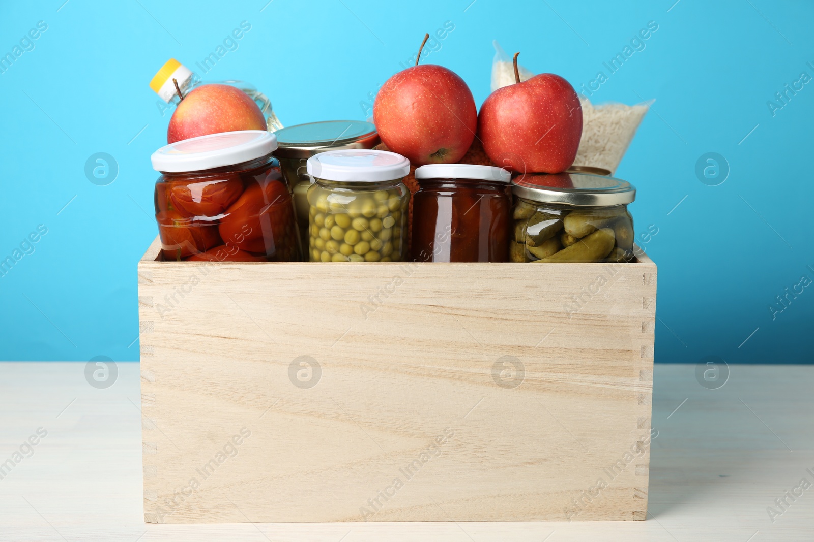 Photo of Different food products for donation in wooden crate on table against light blue background