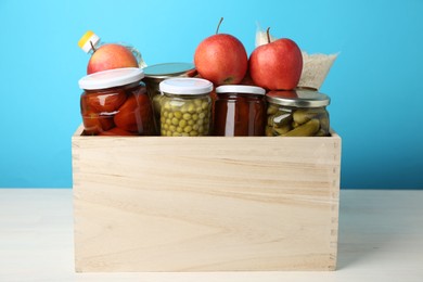 Photo of Different food products for donation in wooden crate on table against light blue background