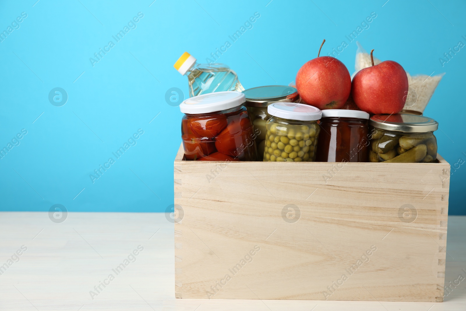 Photo of Different food products for donation in wooden crate on table against light blue background, space for text