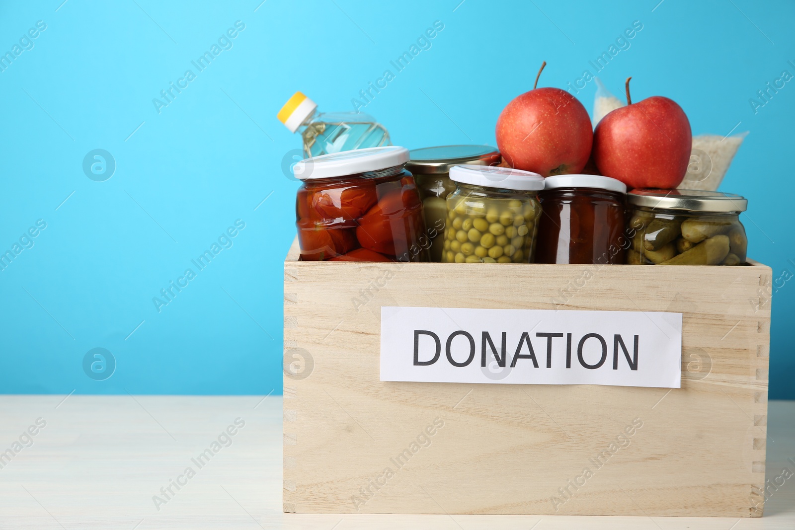 Photo of Different food products for donation in wooden crate on table against light blue background, space for text